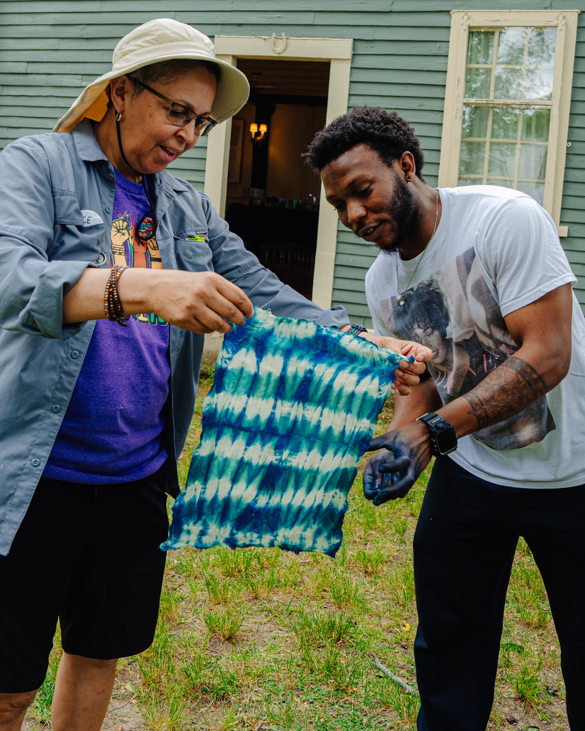 Two people stand outside holding up a blue and white tie-dyed cloth together, examining their work. One person wears a sunhat and glasses; the other has a white T-shirt and blue dye on their hand. A green wooden building is behind them.