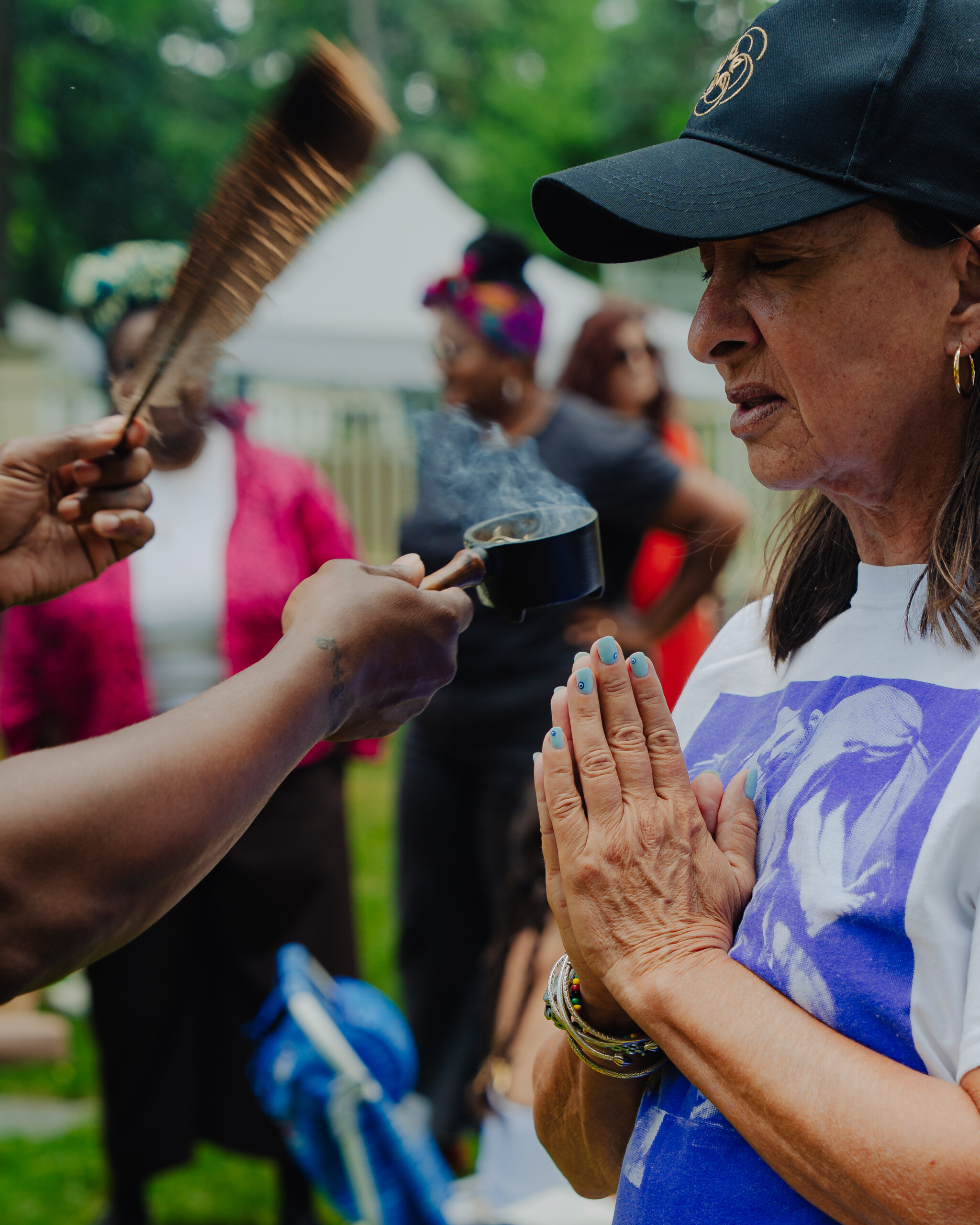 A woman with hands pressed together stands outdoors as another person waves smoke toward her, possibly from a ritual or ceremony. Several people are in the background, and the setting appears to be a gathering or festival.