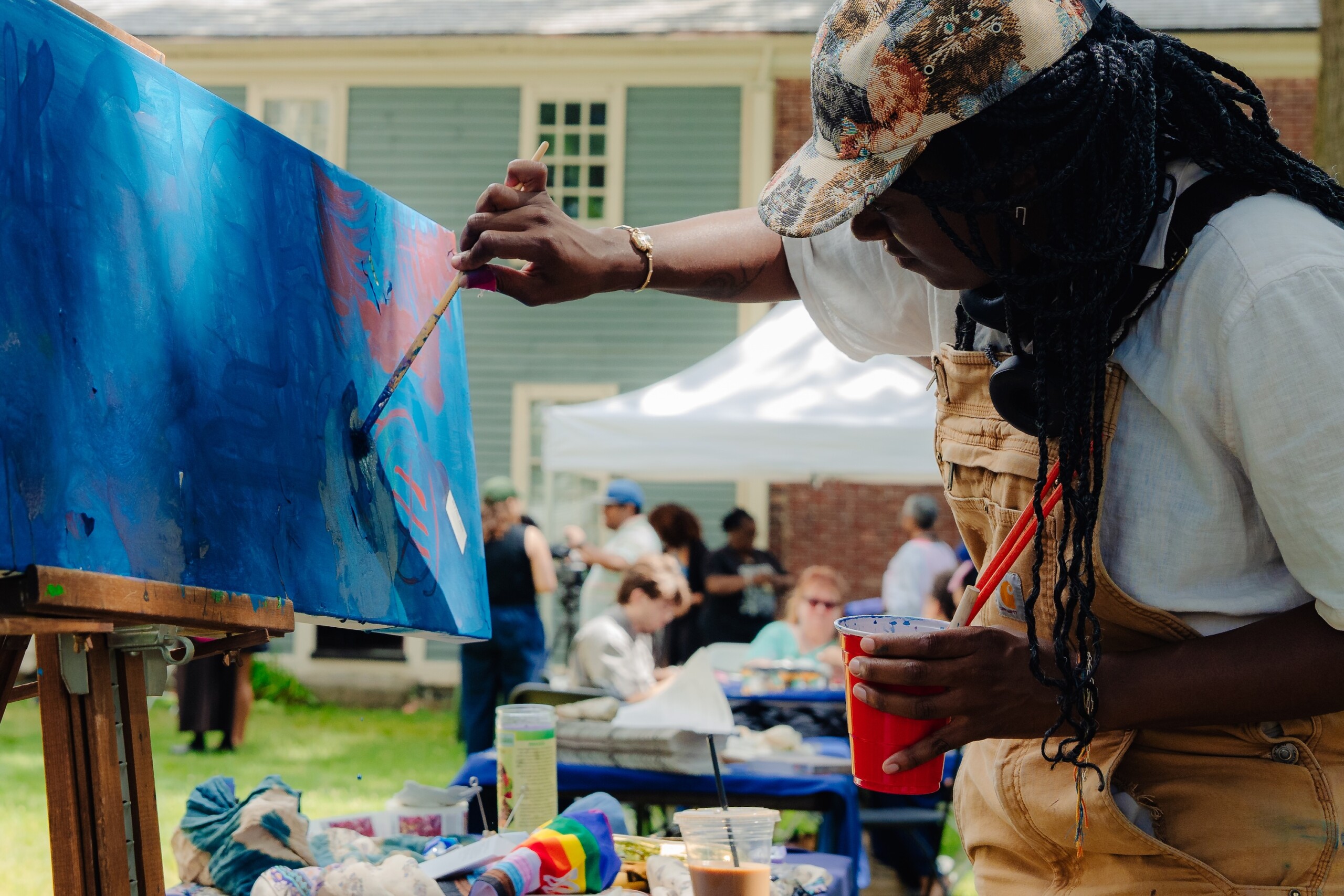 A person wearing a floral cap and overalls paints on a blue canvas at an outdoor event, holding a drink cup, with people and tents visible in the background.