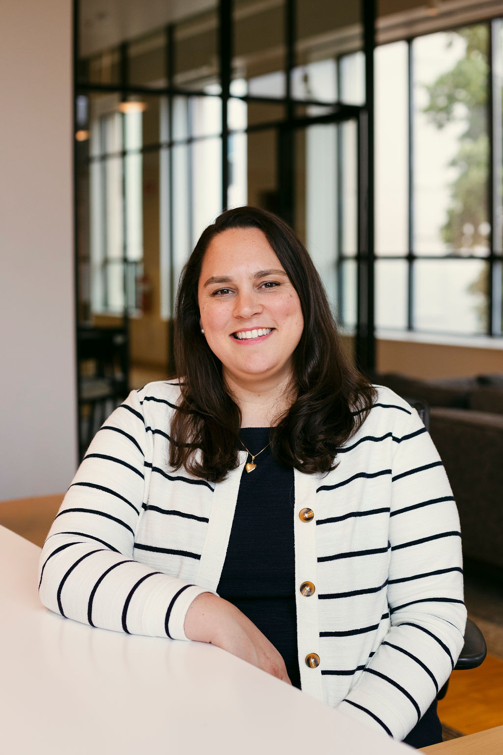 A woman with long brown hair, wearing a white and black striped cardigan over a dark top, sits at a white table and smiles in a modern office with glass walls and natural light.
