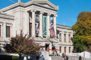 A grand museum building with columns and large banners saying Welcome and displaying artwork hangs on the facade. An American flag is above the steps, and people walk near trees with autumn leaves.