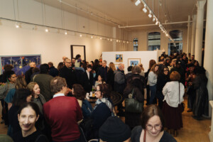 Large crowd of people mingling and viewing art in a brightly lit gallery space with white walls and paintings displayed. The atmosphere is lively and social, with attendees engaged in conversation.