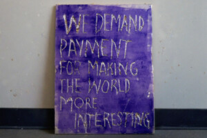A purple sign with rough, hand-painted white letters reads: WE DEMAND PAYMENT FOR MAKING THE WORLD MORE INTERESTING. The sign is propped against a plain wall.