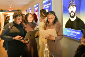 A group of women stand in a hallway, smiling and working on laptops. Behind them, posters featuring award recipients, including one labeled Innovation Leader Award, are displayed on the wall.