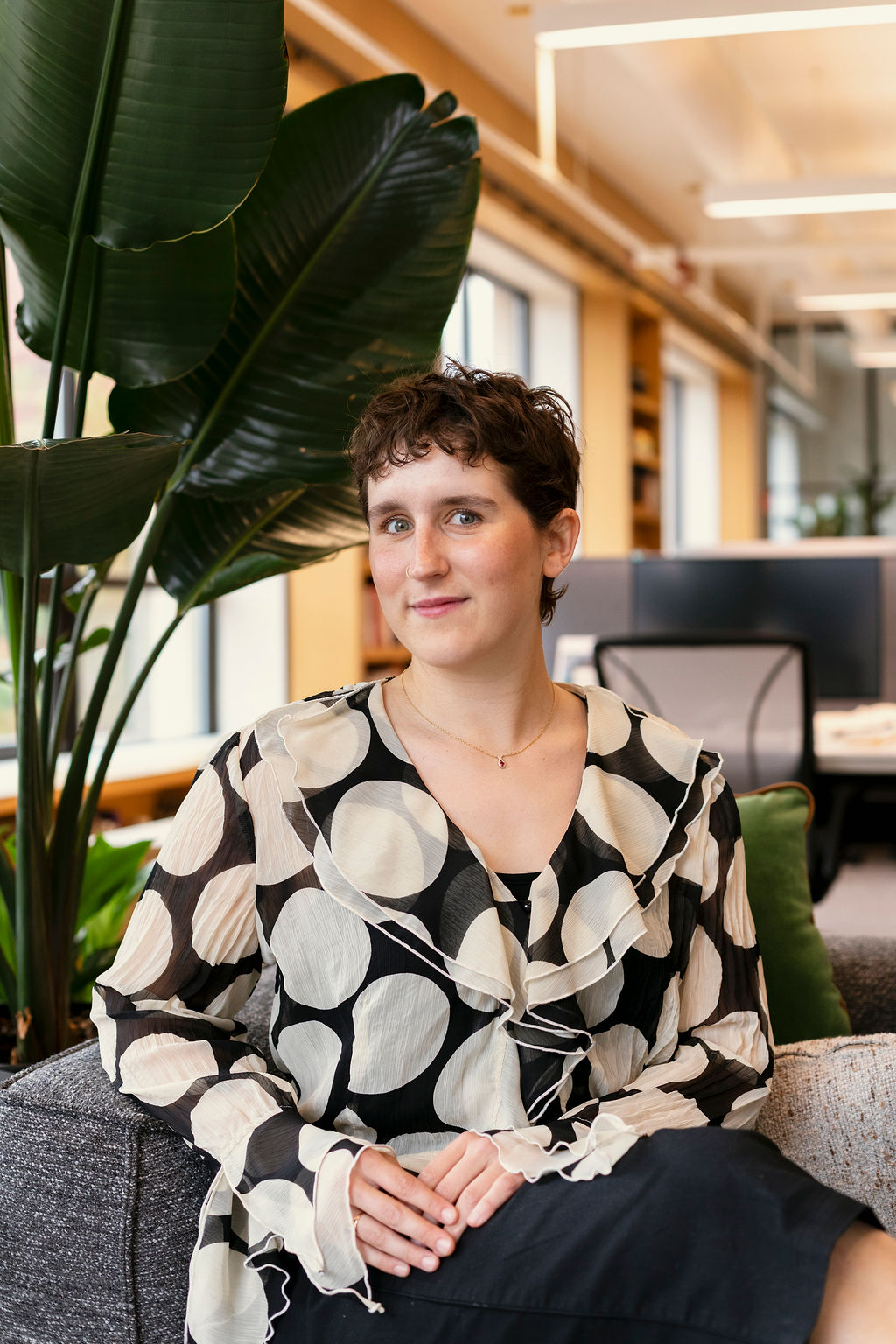 A person with short curly hair sits on a gray couch in a modern office, wearing a black and white polka dot blouse. Large green plants and desks are visible in the background.