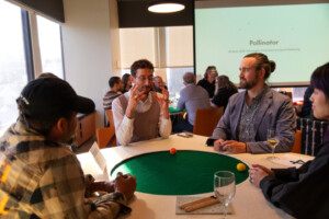 A group of people sit around a green circular table, engaged in conversation, with drinks in front of them. A presentation screen in the background reads Pollinator. The setting appears to be a social or networking event.