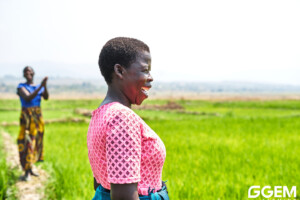 A smiling woman in a pink top stands in a lush green field, with another woman in colorful clothing clapping in the background. The sky is bright and the atmosphere appears joyful. GGEM logo is visible in the corner.