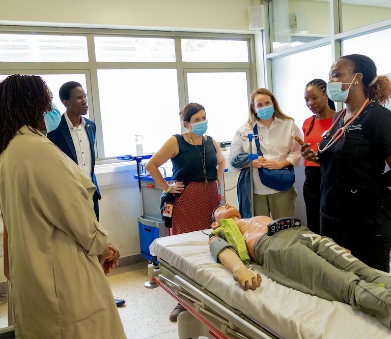 A group of people wearing masks stands around a medical mannequin on a hospital bed, listening to a healthcare professional in a clinical setting.