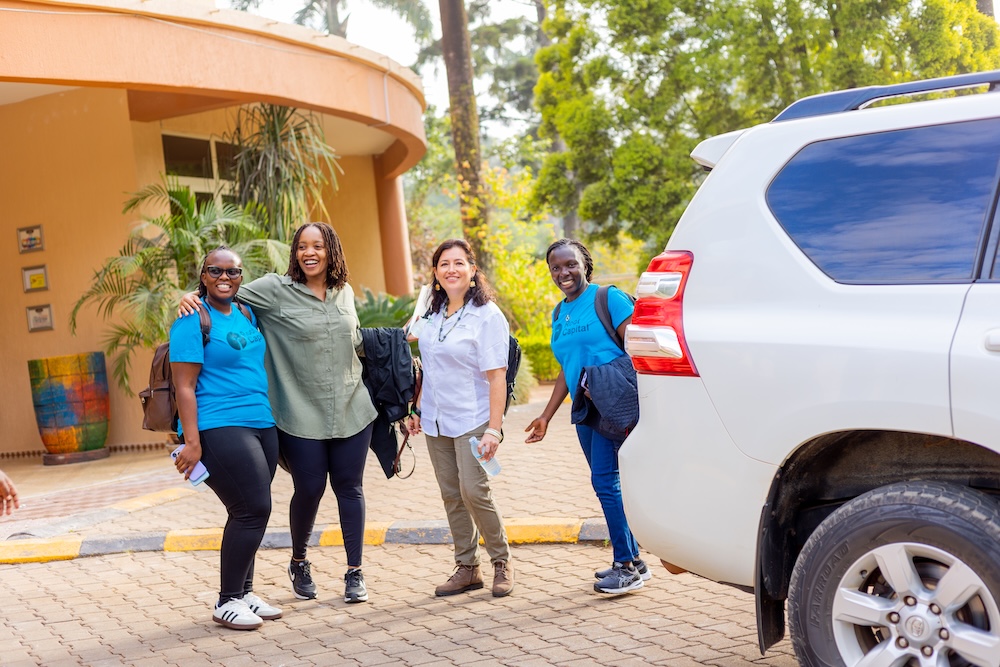 Four smiling women stand together on a paved area next to a white SUV, with trees and a building in the background. Two women wear blue shirts, one wears green, and one wears white.