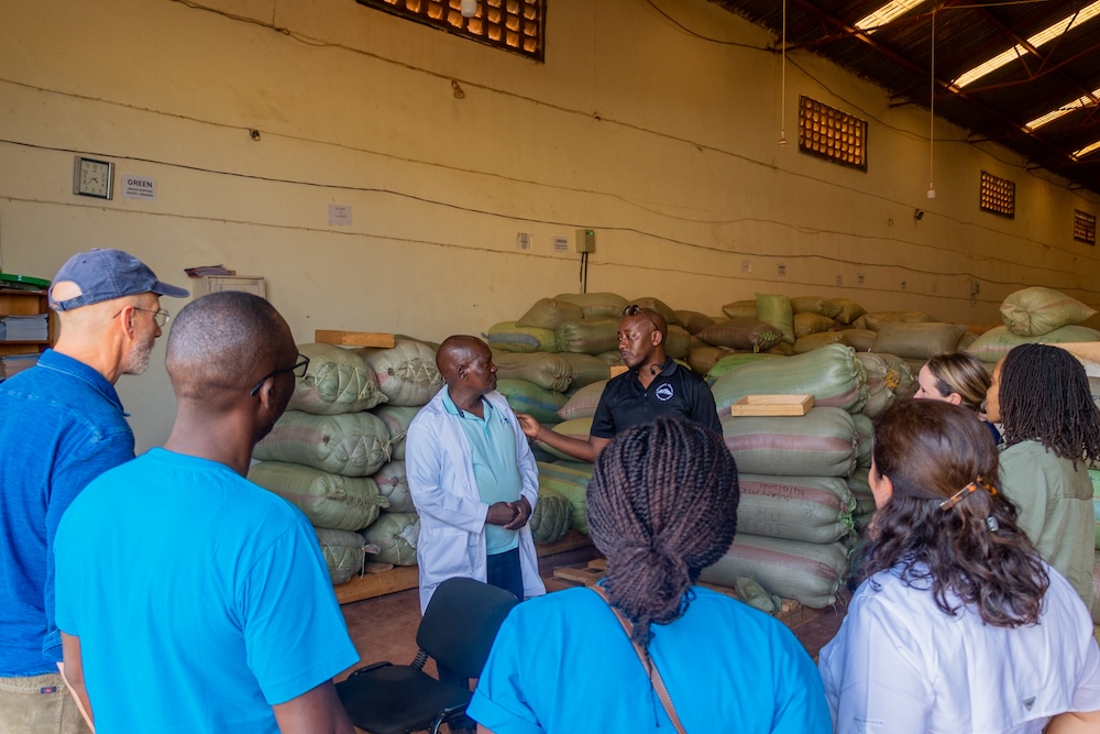 A group of people listens to two men speaking in a warehouse filled with large sacks stacked against the walls.