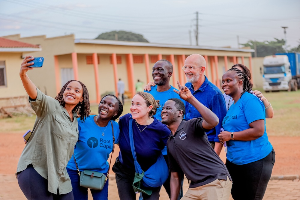 A group of seven people, smiling and posing for a selfie outdoors by a building. Some wear blue shirts with Root Capital text. It is daytime, and the mood is cheerful.