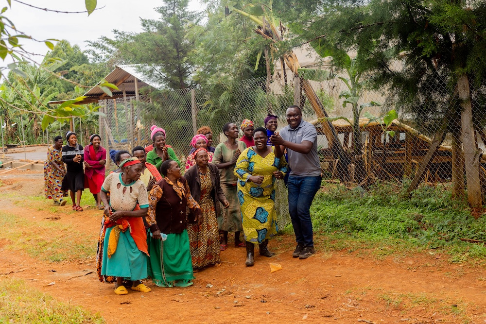 A group of women and one man smile and walk together on a dirt path outdoors, with trees, plants, and a wire fence in the background.