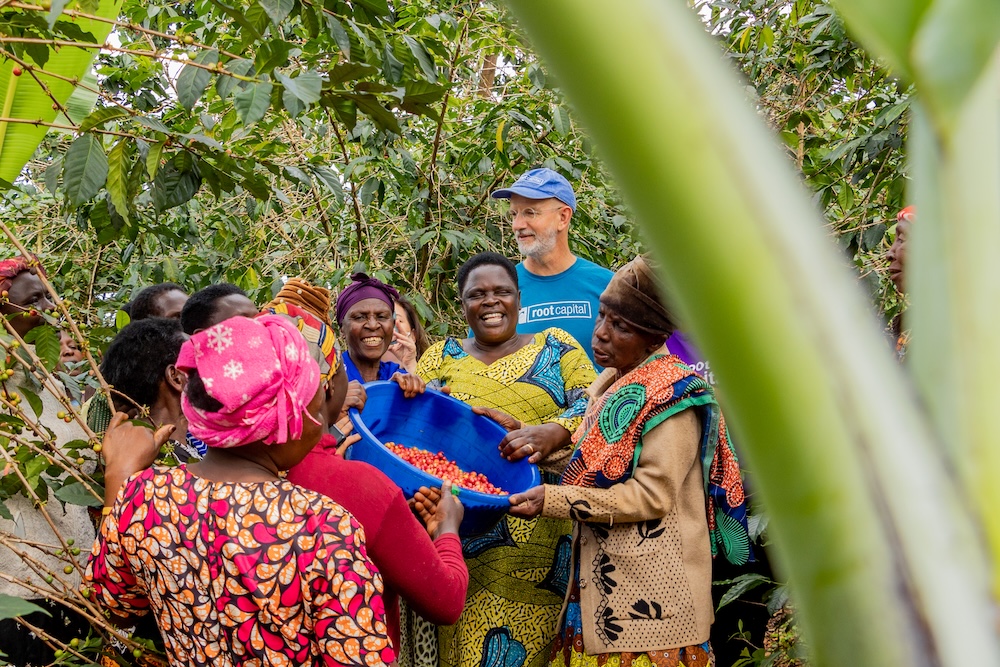 A group of people, mostly women, gather among green plants. One woman in colorful attire holds a blue tub of red berries. Everyone is smiling and interacting happily. A man in a blue shirt and cap stands behind them.