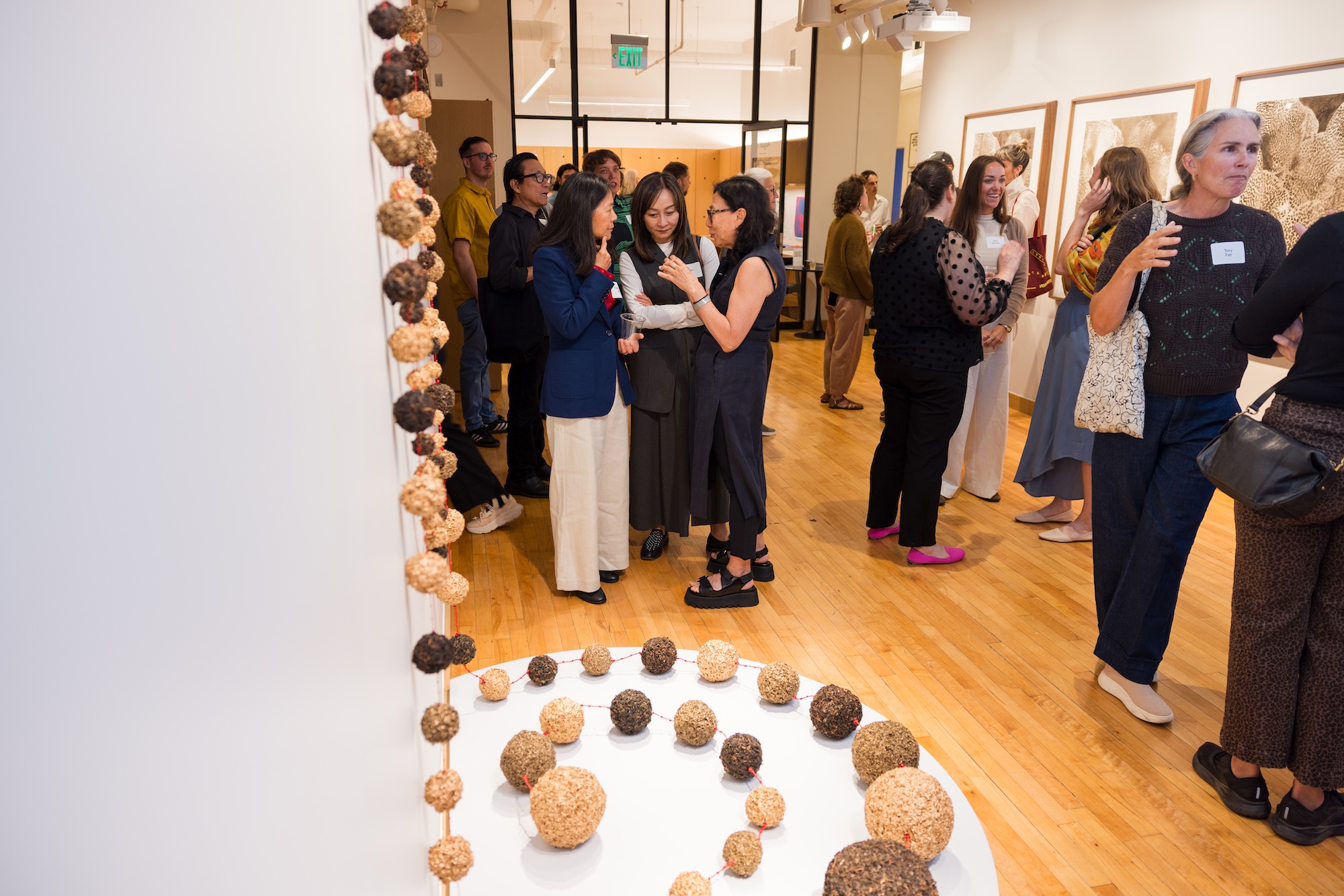 People gather in an art gallery, chatting and observing exhibits. Spherical sculptures are displayed on a white table in the foreground. Framed artwork hangs on the walls, and the room is brightly lit with wooden floors.