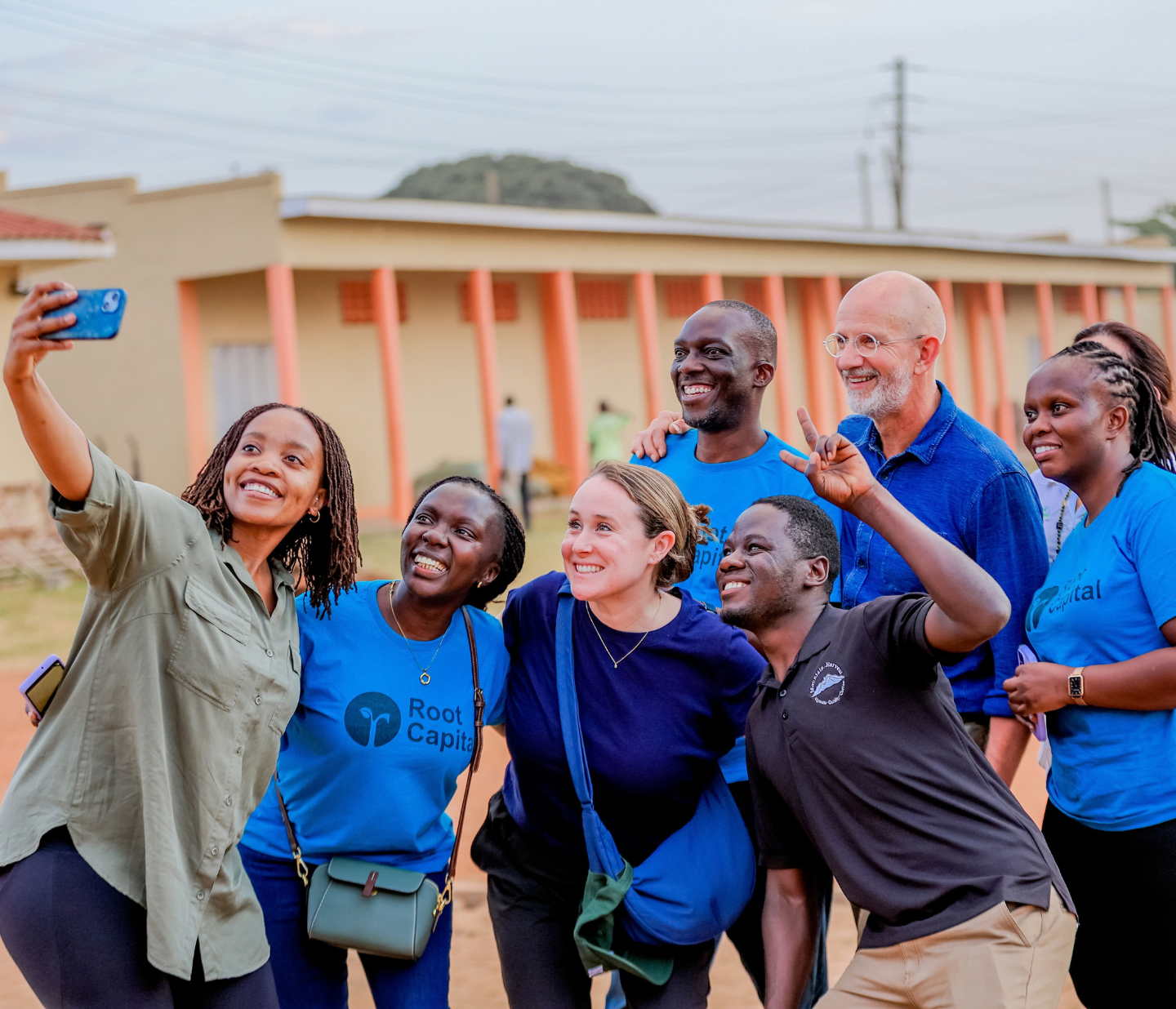 A group of seven people, some wearing Root Capital shirts, gather and smile for a selfie outdoors, in front of a building with orange columns.