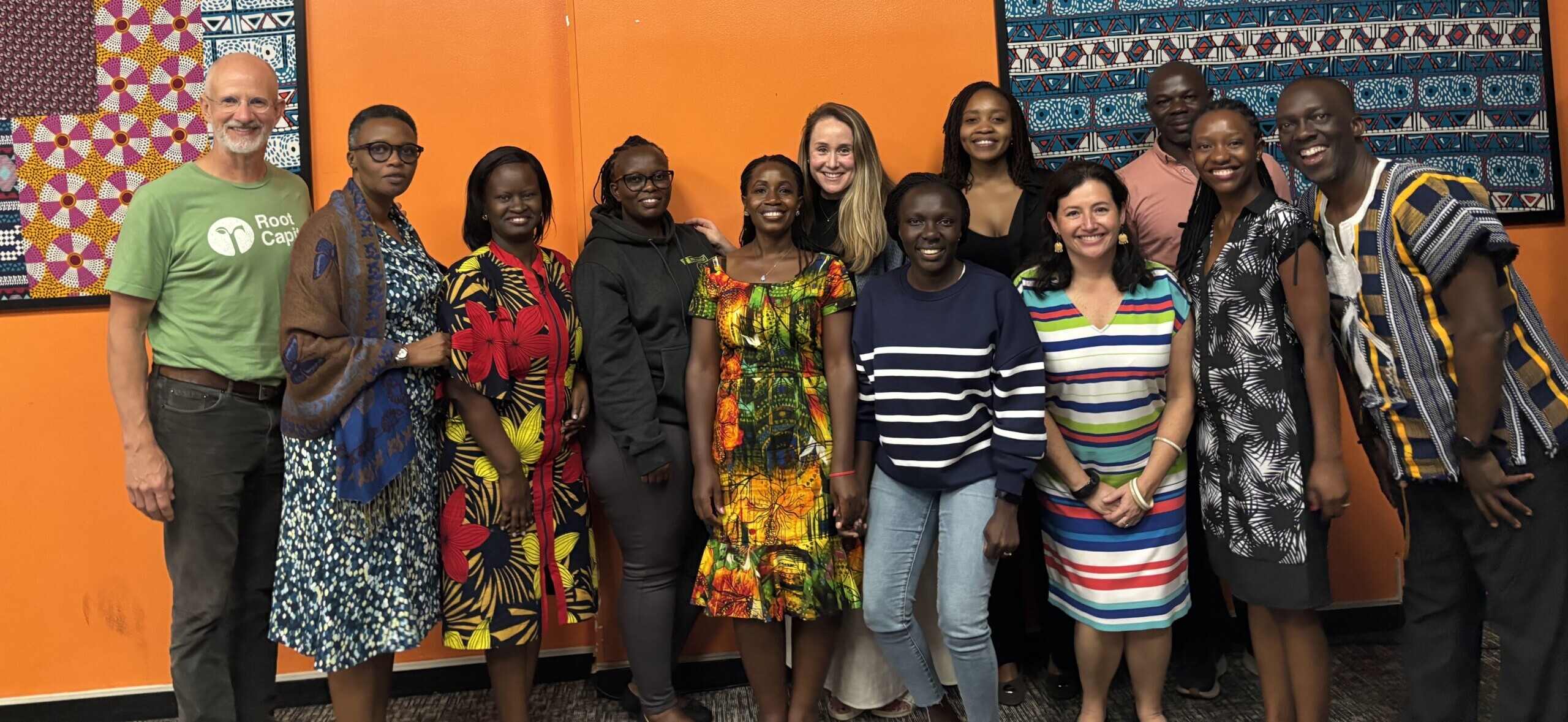 A group of thirteen people stand smiling in front of a bright orange wall decorated with colorful patterned textiles. They appear happy and relaxed, posing together for a group photo indoors.